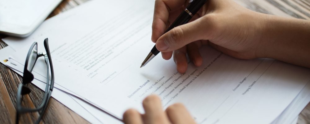 Unrecognizable businessman examining papers at table. Manager with ballpoint pen filling business papers. Close-up of male hands working at desk. Analyzing documents concept